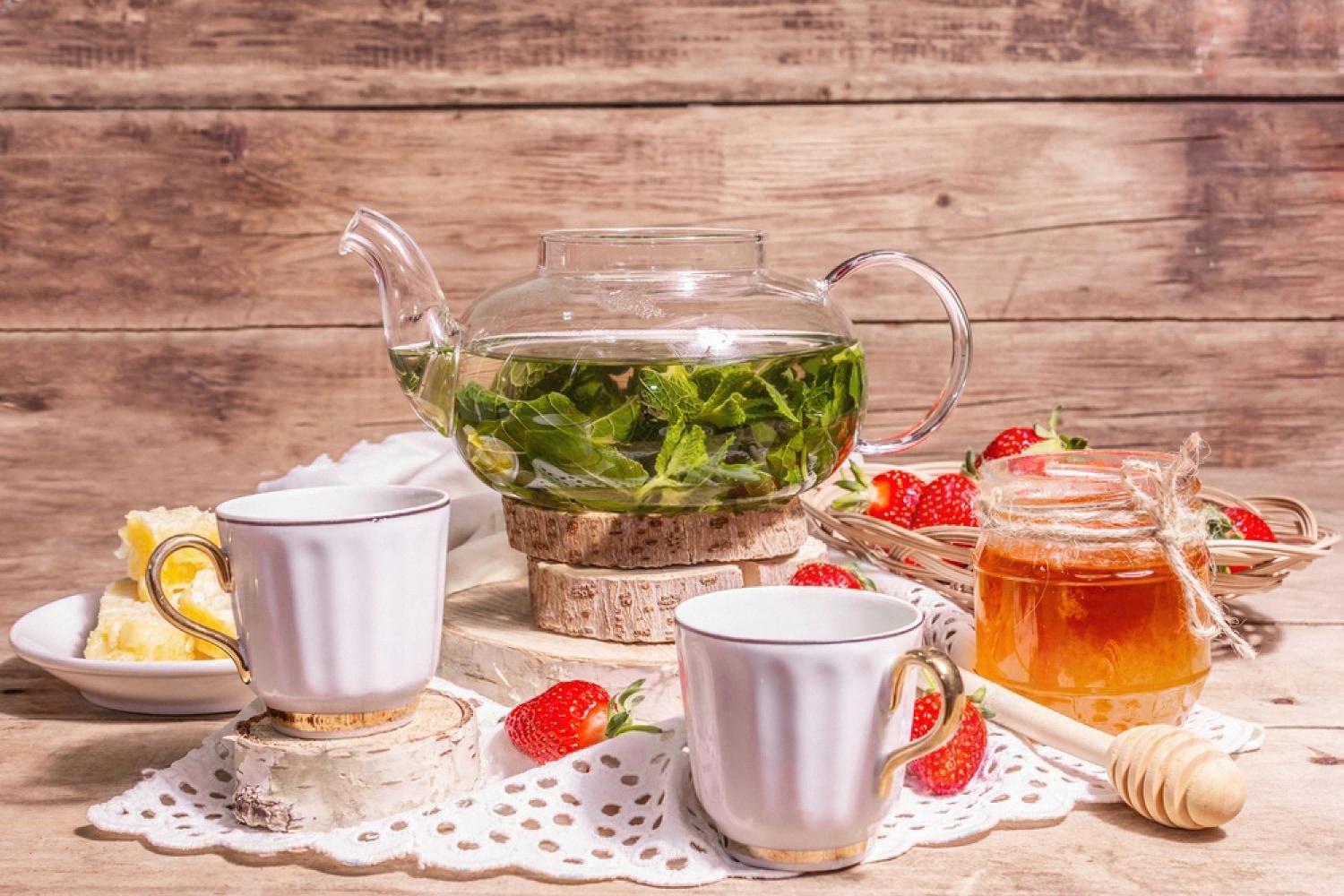 Glass teapot with fresh mint leaves, two porcelain cups, strawberries and honey on a rustic table